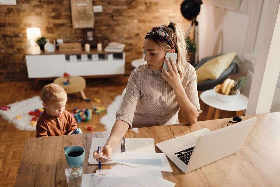 אישה העובדת מהבית בטלפון מול מחשב נייד וילד קטן יושב לצידה , GettyImages